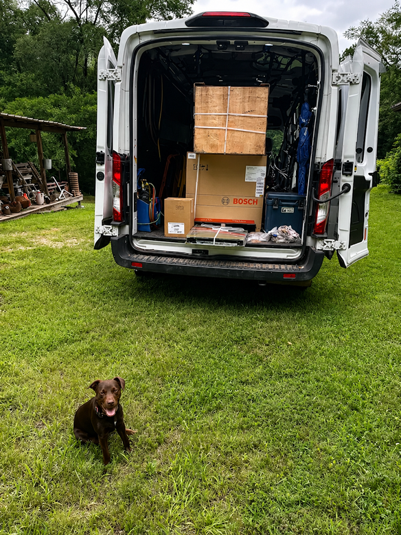 White van with open rear doors loaded with boxes on a grassy yard, with a small dog in the foreground.