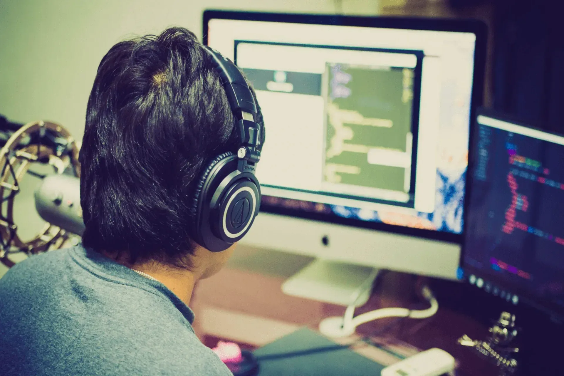 Person wearing headphones works on computer, focused, indoors. Second monitor shows data, desk set up.