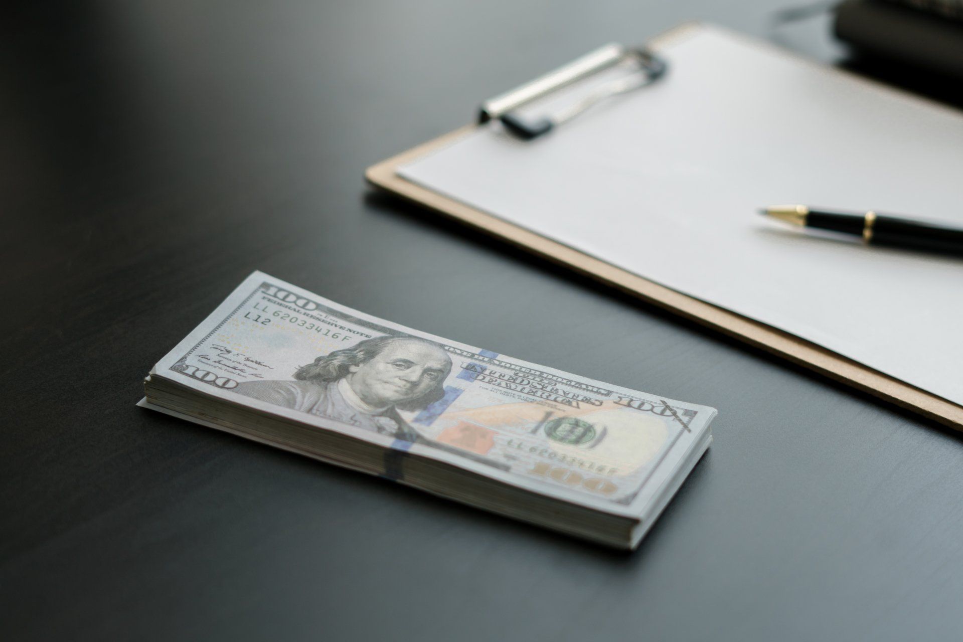 Stack of US $100 bills beside a clipboard with blank paper and a pen on a dark surface.