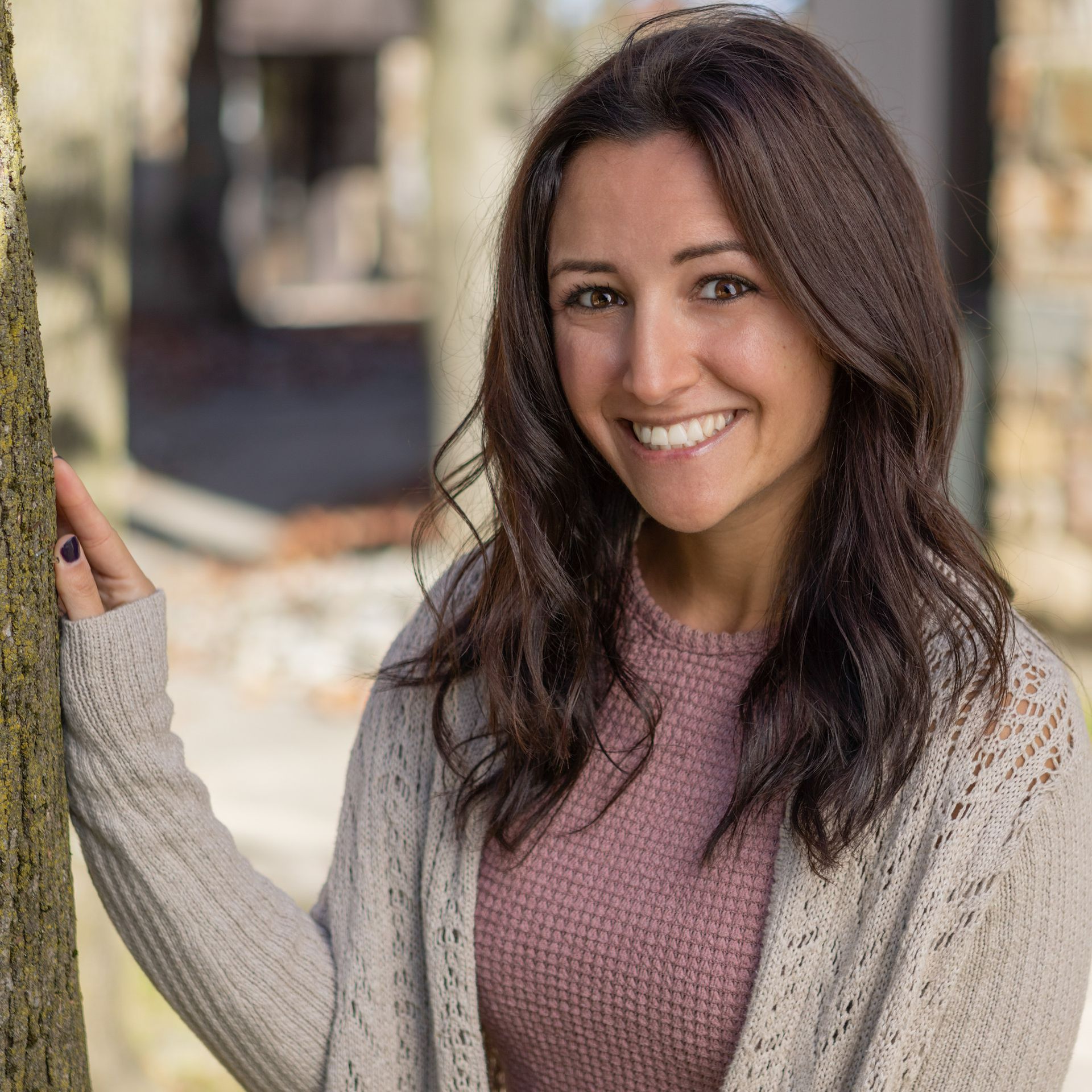 A woman in a sweater is leaning against a tree and smiling.