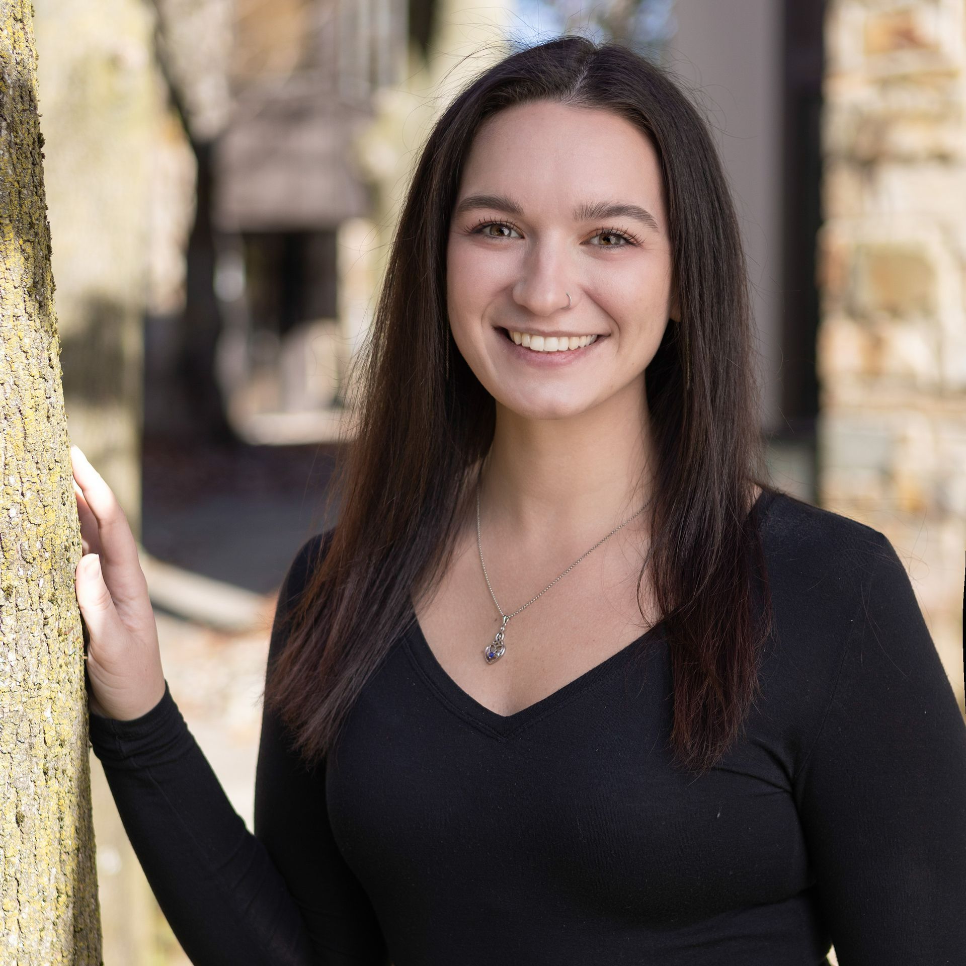 A woman in a black shirt is leaning against a tree and smiling.