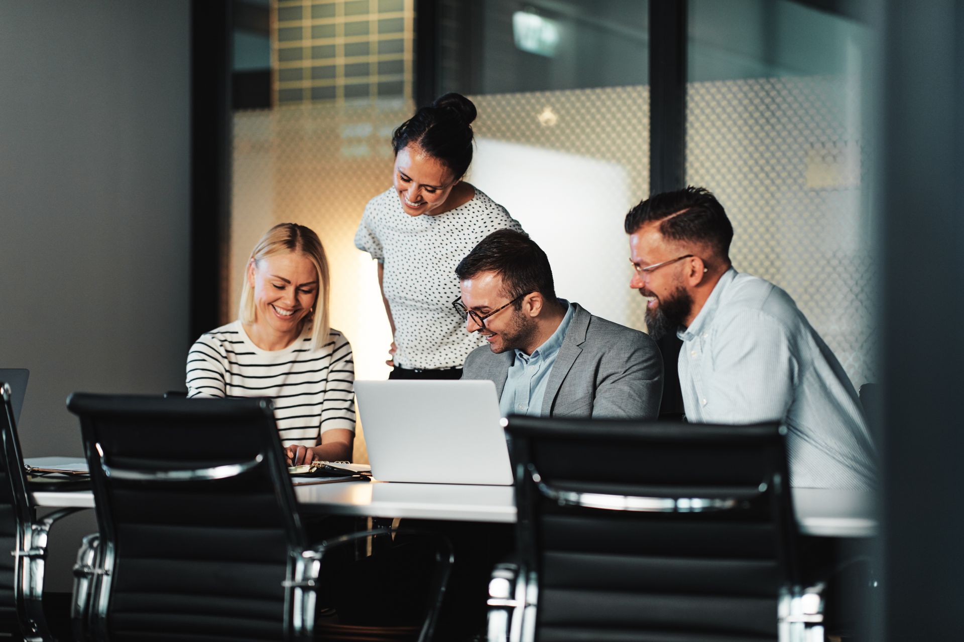 Four professionals collaborate at a table in a modern office, looking at a laptop screen and smiling.