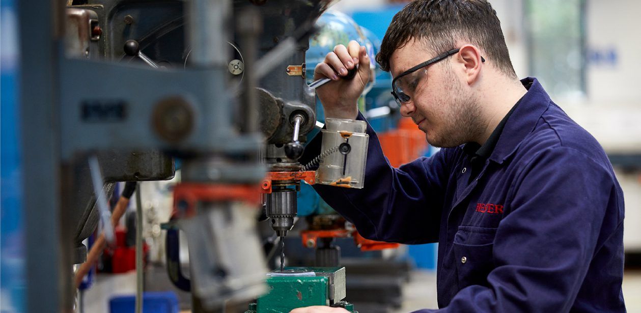 A person in blue coveralls and safety glasses adjusts a metal drilling machine in a workshop.