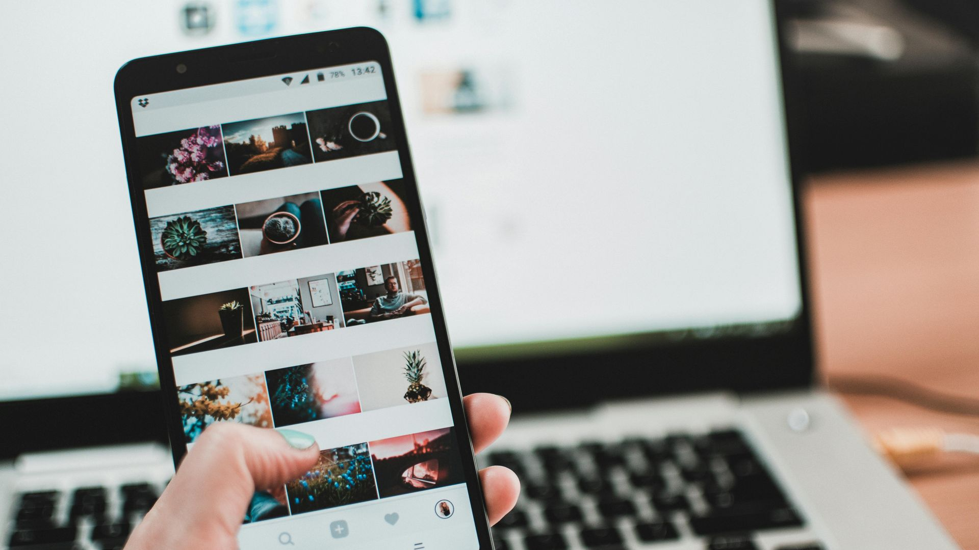 A person holds a smartphone displaying a grid of photos on a social media app, with a laptop blurred in the background.