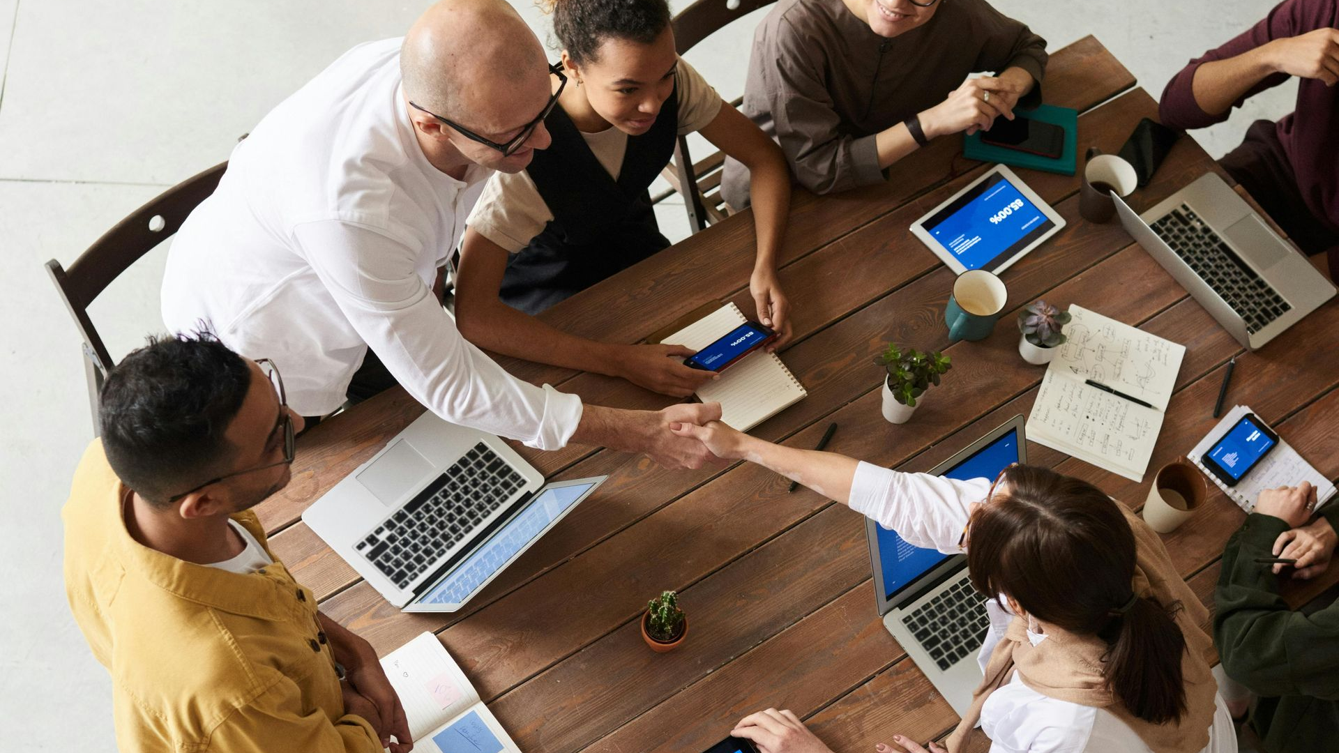 A high-angle shot of a diverse group of people around a wooden table, shaking hands during a collaborative office meeting.