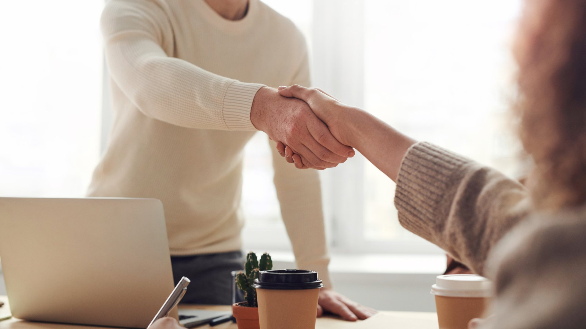 Two people shake hands over a desk with a laptop, a notebook, and two coffee cups in a bright office.