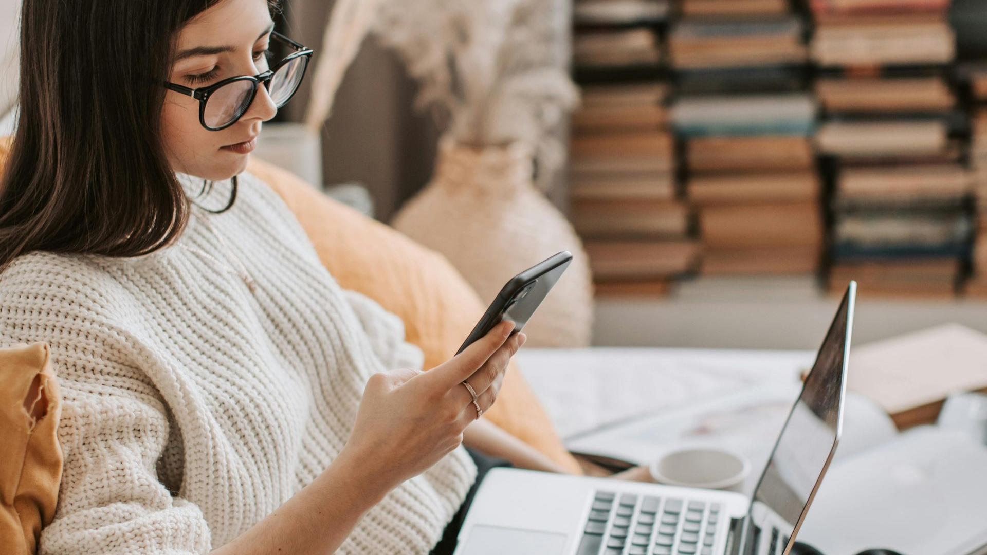 A person wearing glasses and a cream sweater sits at a desk, looking at a smartphone with a laptop open nearby.