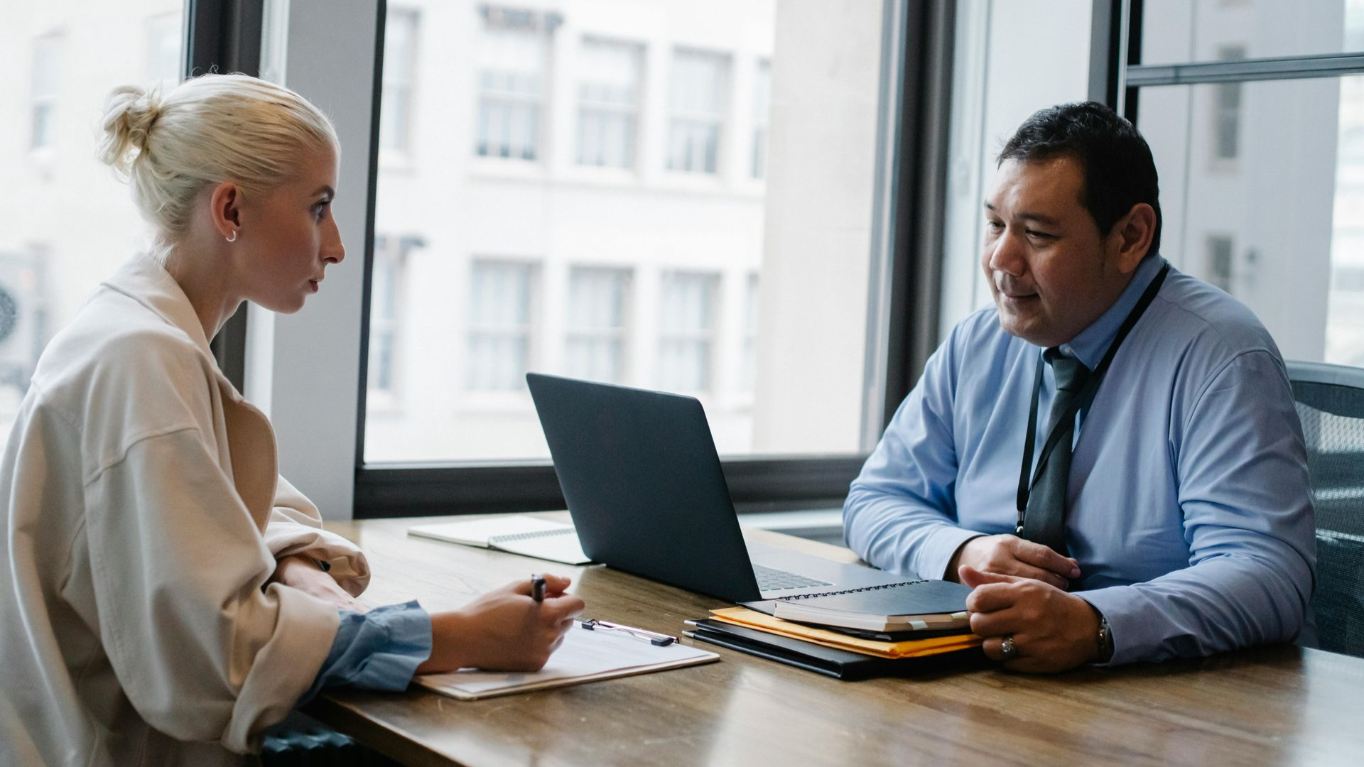 A person in a light-colored blazer interviews with a person in a blue shirt and tie at a table with a laptop.