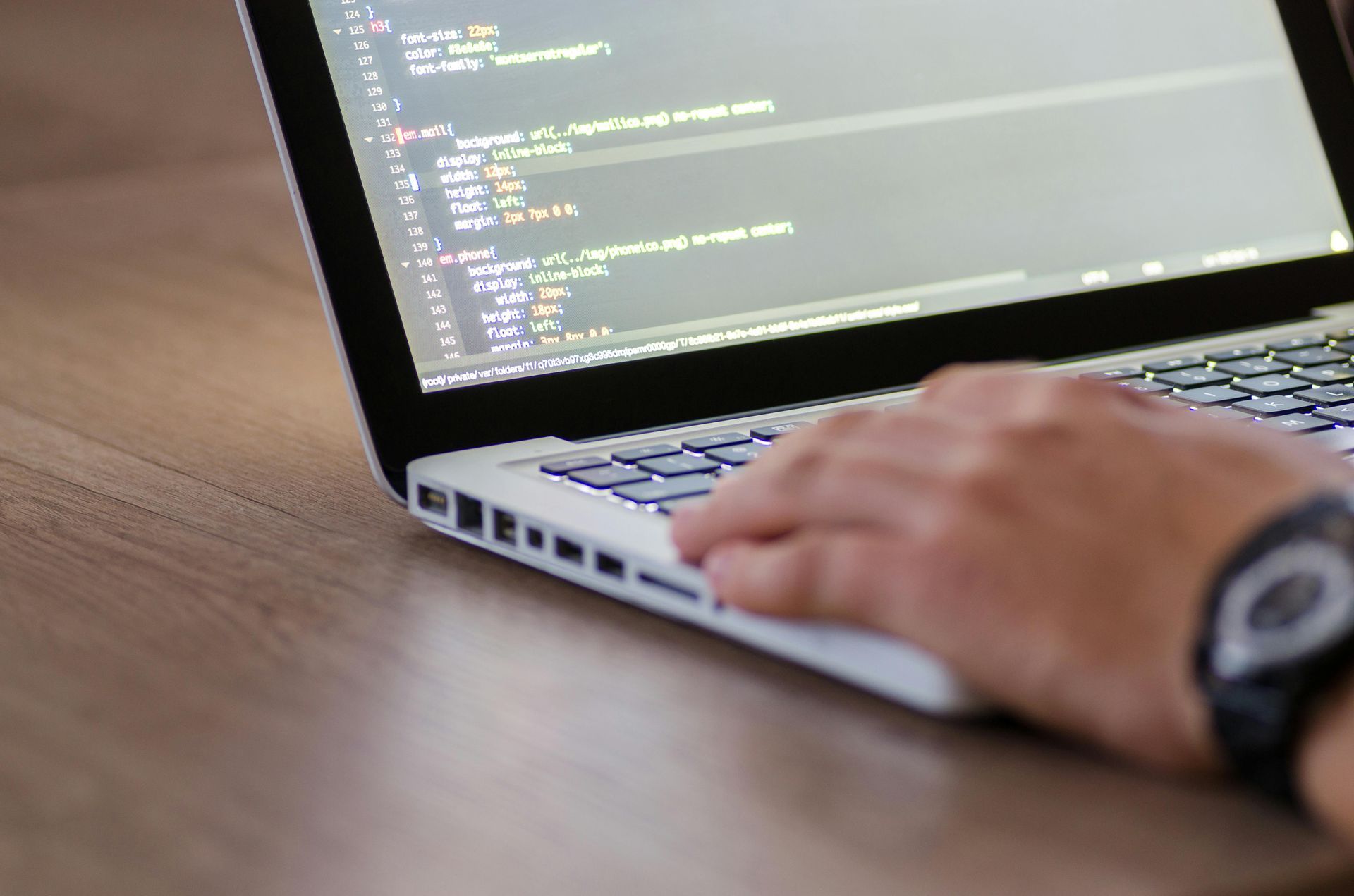 Close-up of a person typing code on a laptop screen while working at a wooden desk.