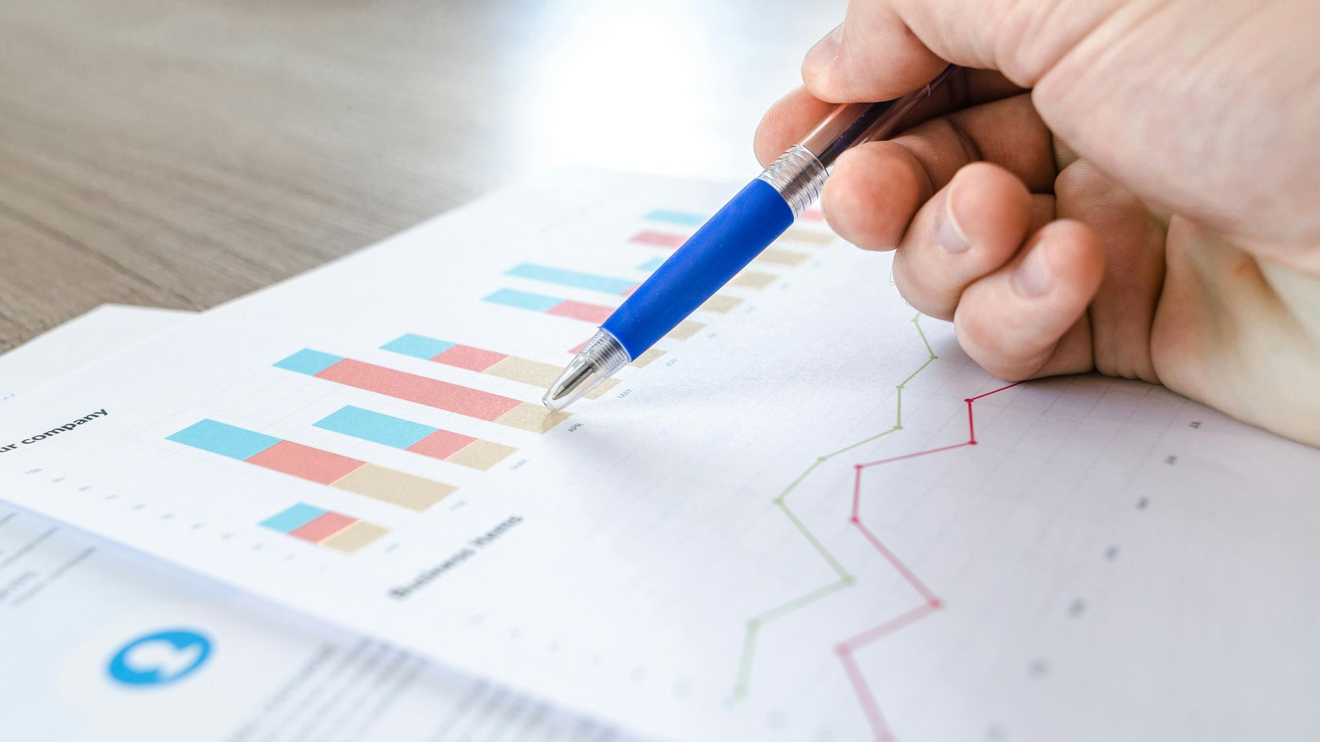 A hand holds a blue pen pointing at a paper featuring stacked bar charts and line graphs on a wooden surface.