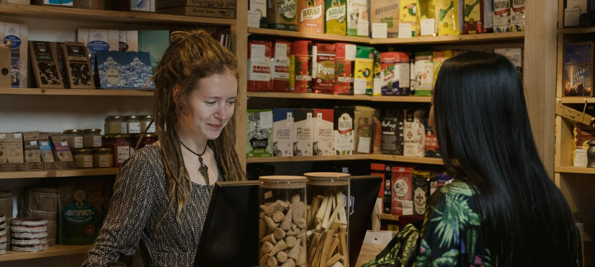 Two people interact at a shop counter, with a clerk smiling while facing a customer in a store with shelves of products.