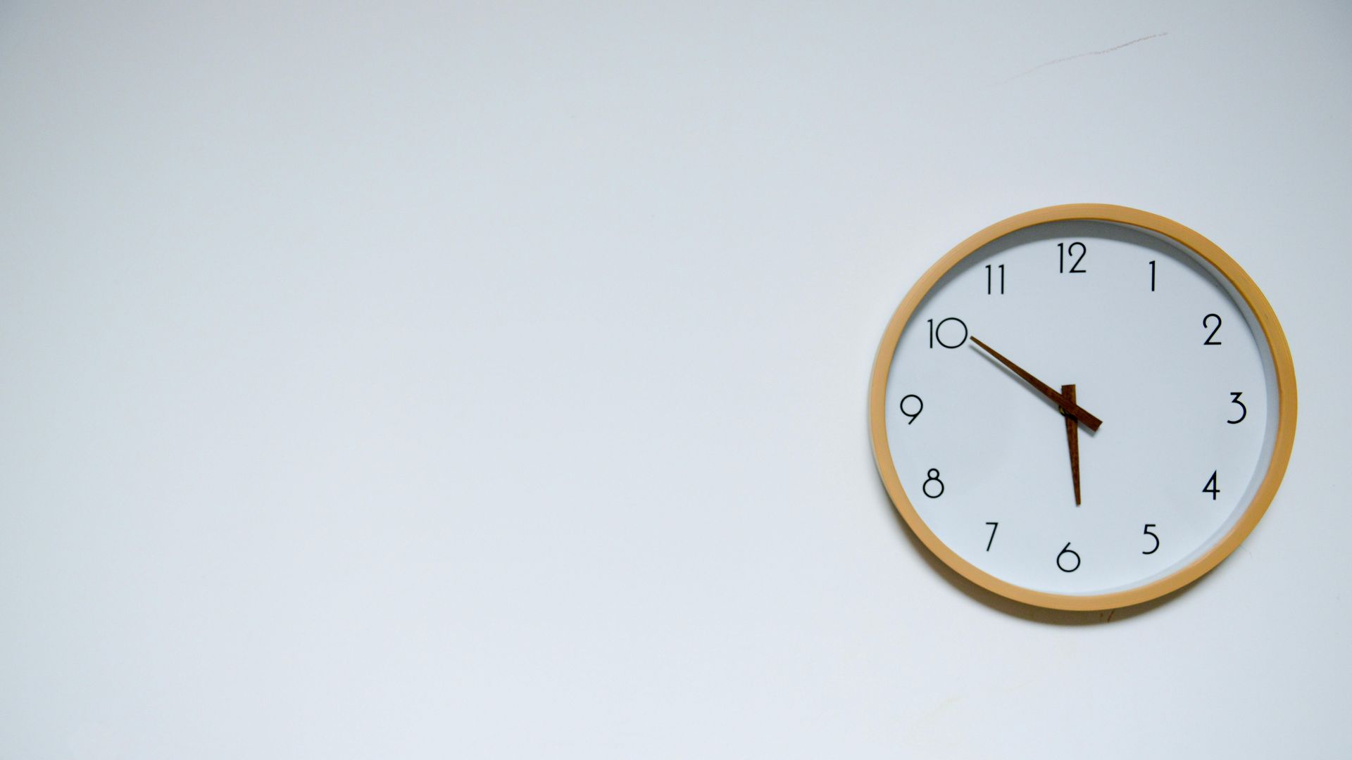 A light wood-framed analog wall clock on a plain white wall, showing the time as approximately 4:50.