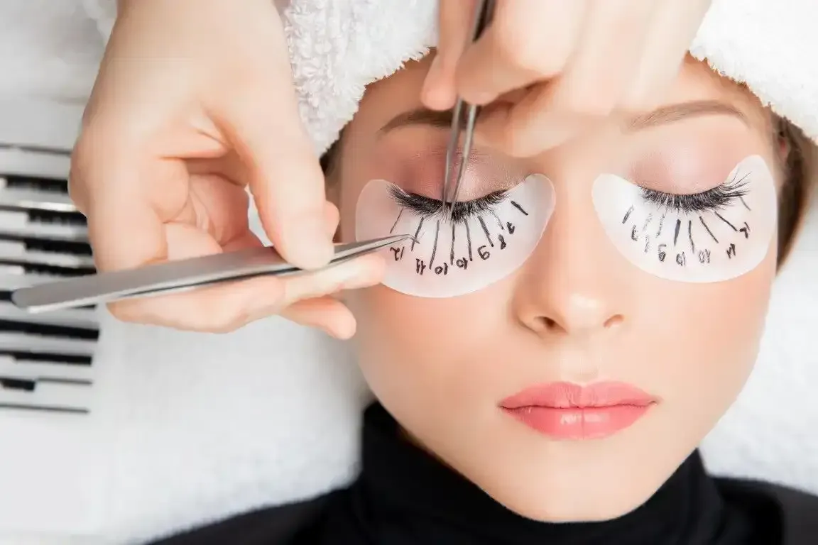 Eyelash extensions being applied; woman lying down with eye patches and tweezers.