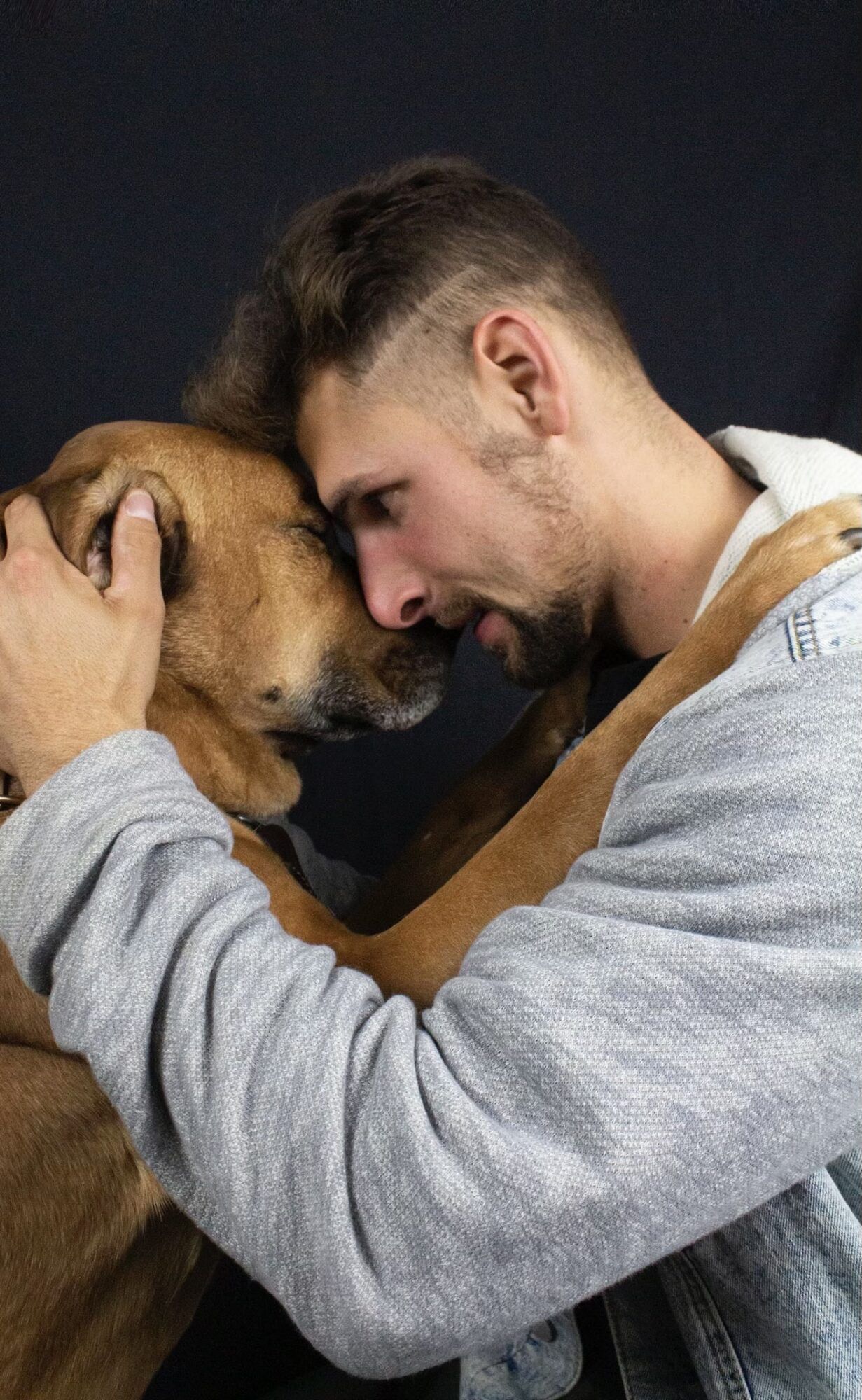 A man is hugging a brown dog with his head on the dog 's head.