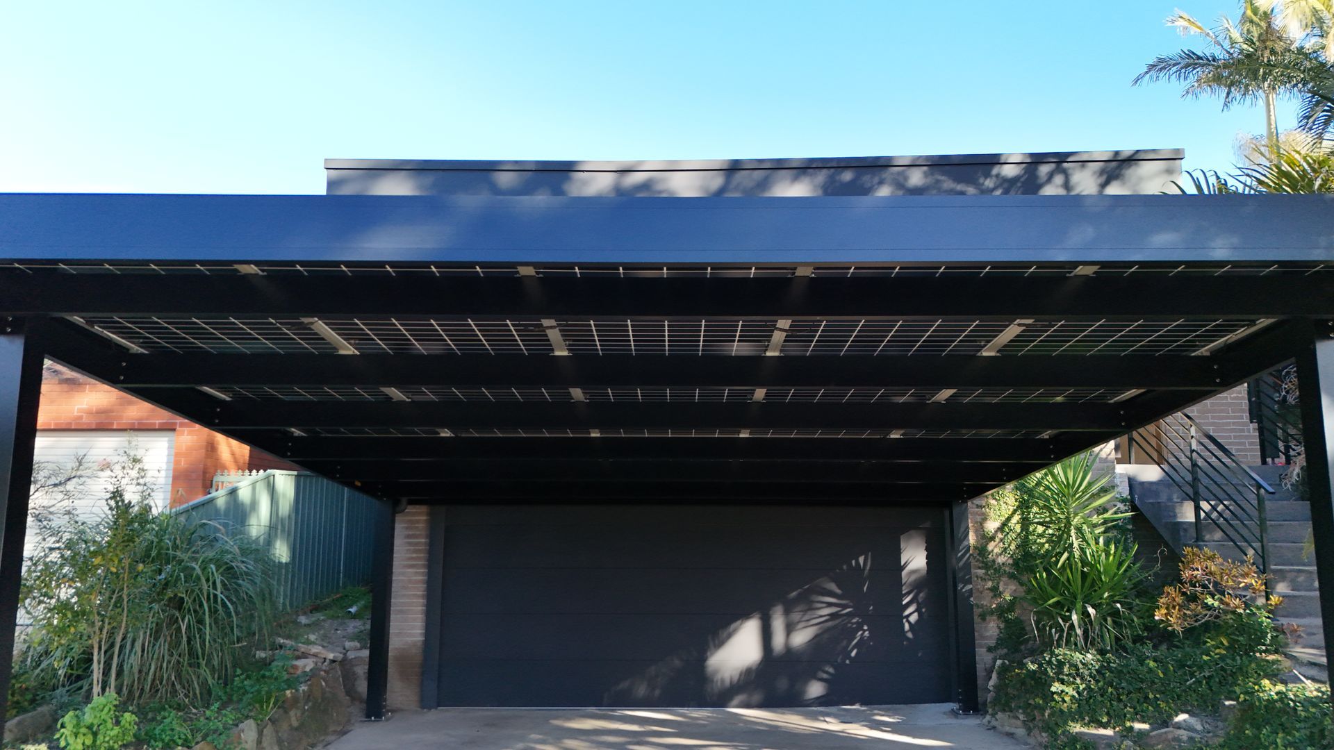 Black Carport With Solar Panels Over a Garage Door, Plants in Front — Mid North Coast Solar Carports in Coffs Harbour, NSW