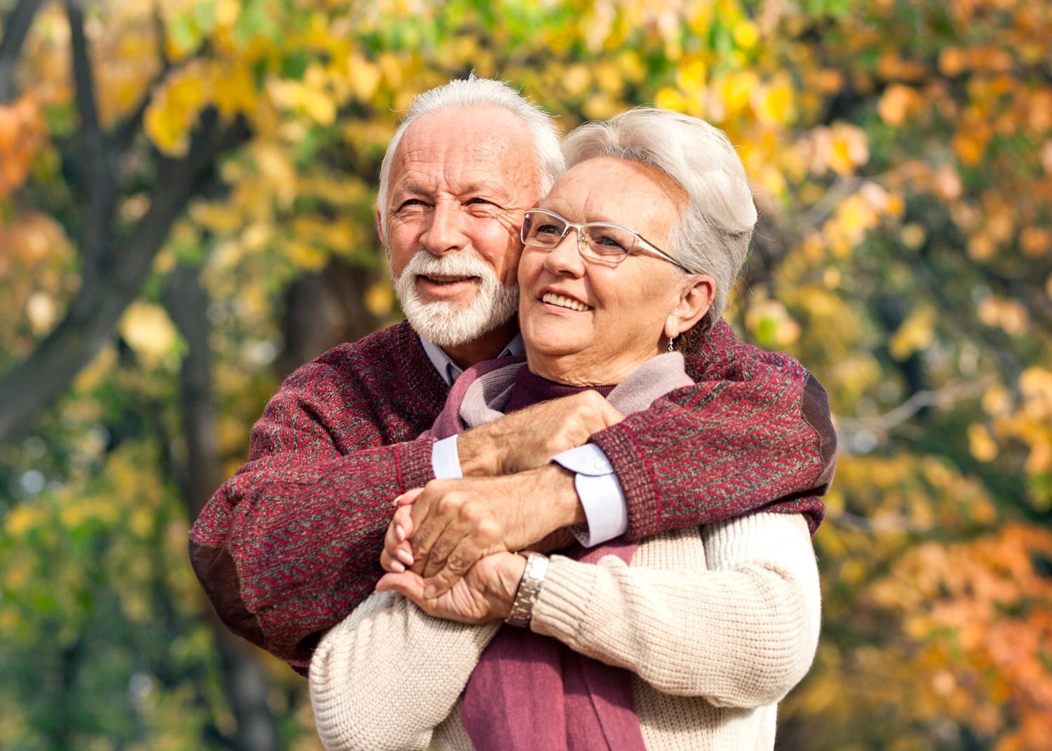 Older couple embraces, smiling outdoors in autumn with colorful foliage.