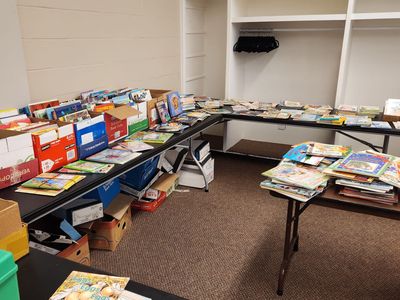 Books displayed on tables and in boxes; likely a book sale. Beige walls and carpet.