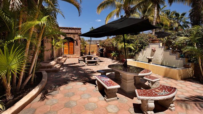 Sunny patio with stone tables and benches, palm trees, and a black umbrella outside a rustic building
