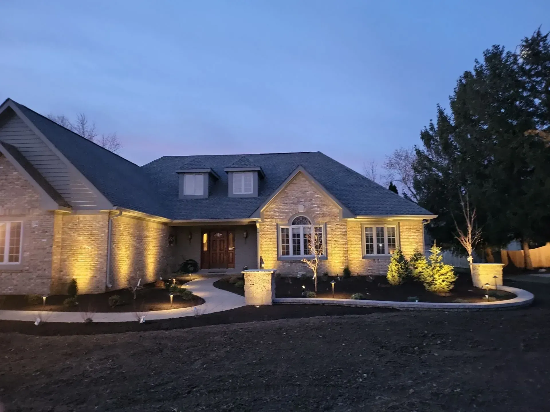 House exterior at dusk, lit with spotlights, features brick facade, dark roof, and landscaped yard.