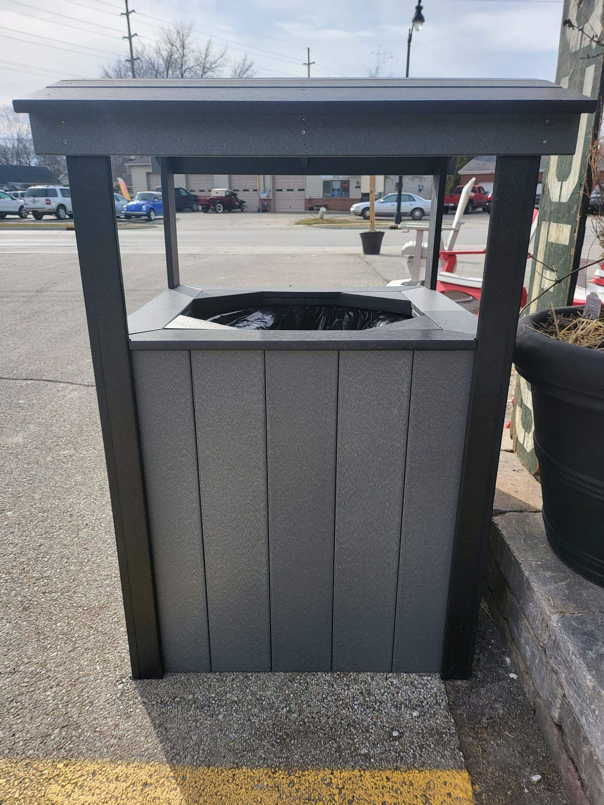 A trash can is sitting on the side of the road in a parking lot.