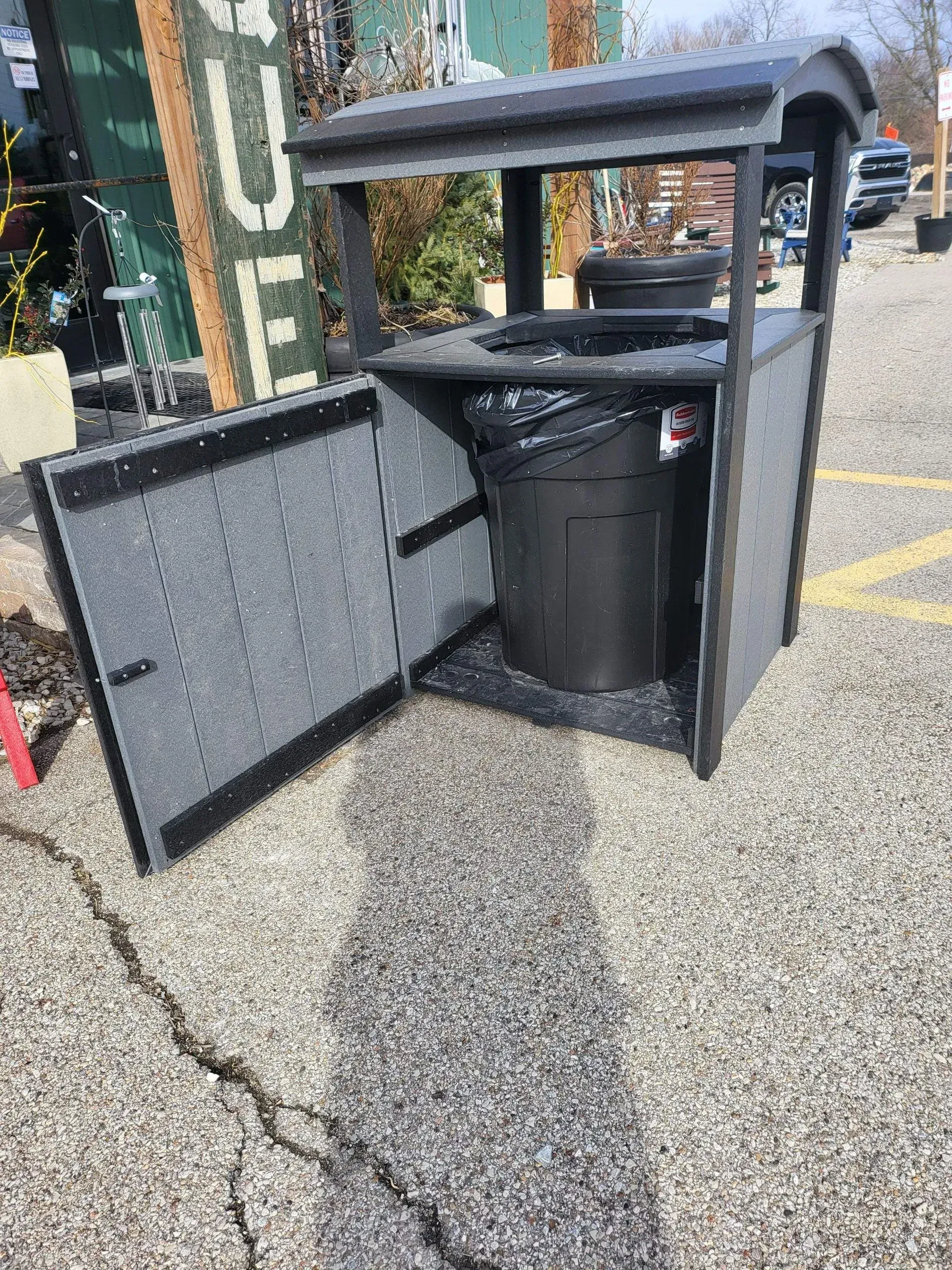 A trash can is sitting on the side of the road in a parking lot.