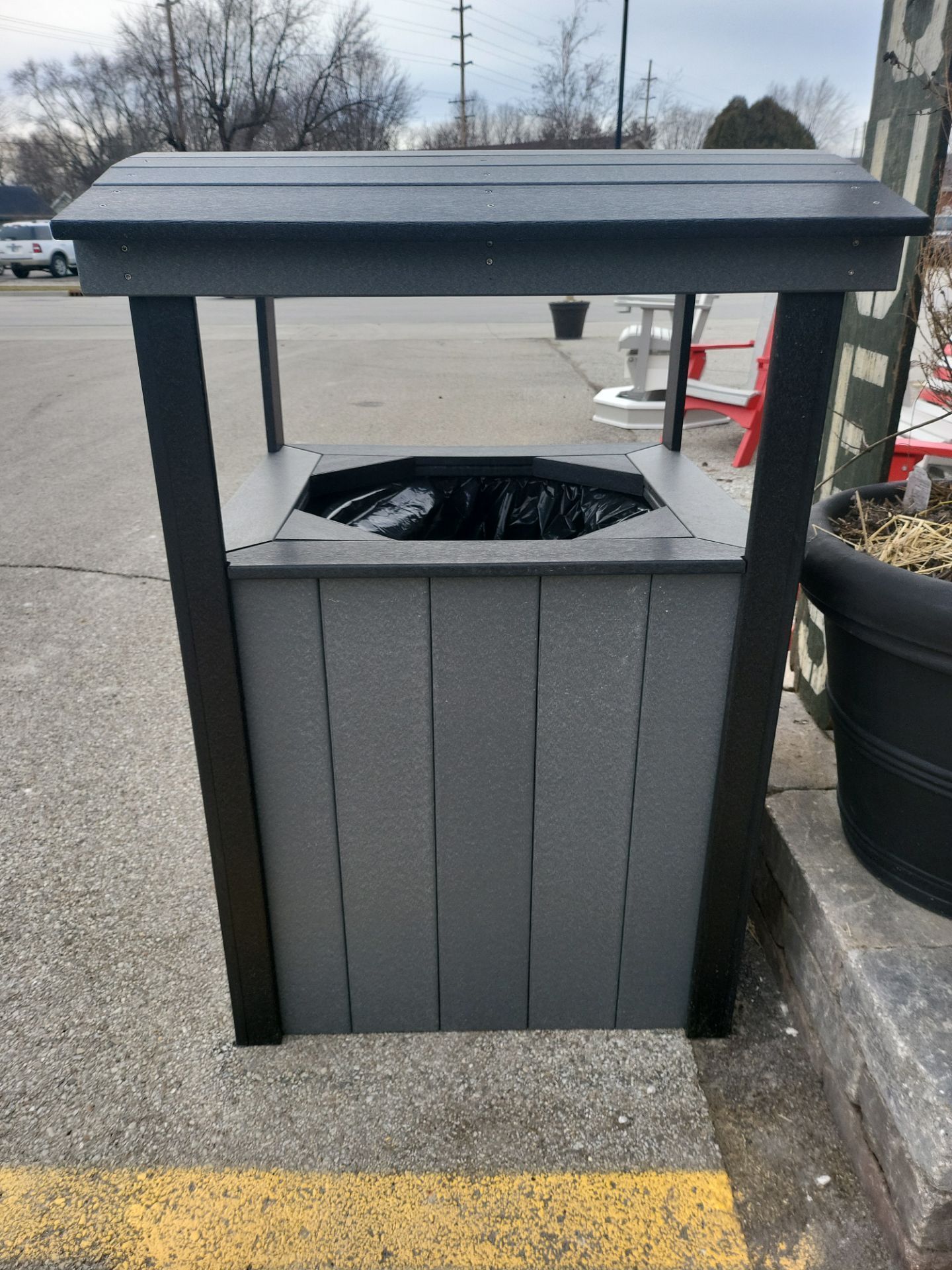 A trash can is sitting on the side of the road in a parking lot.