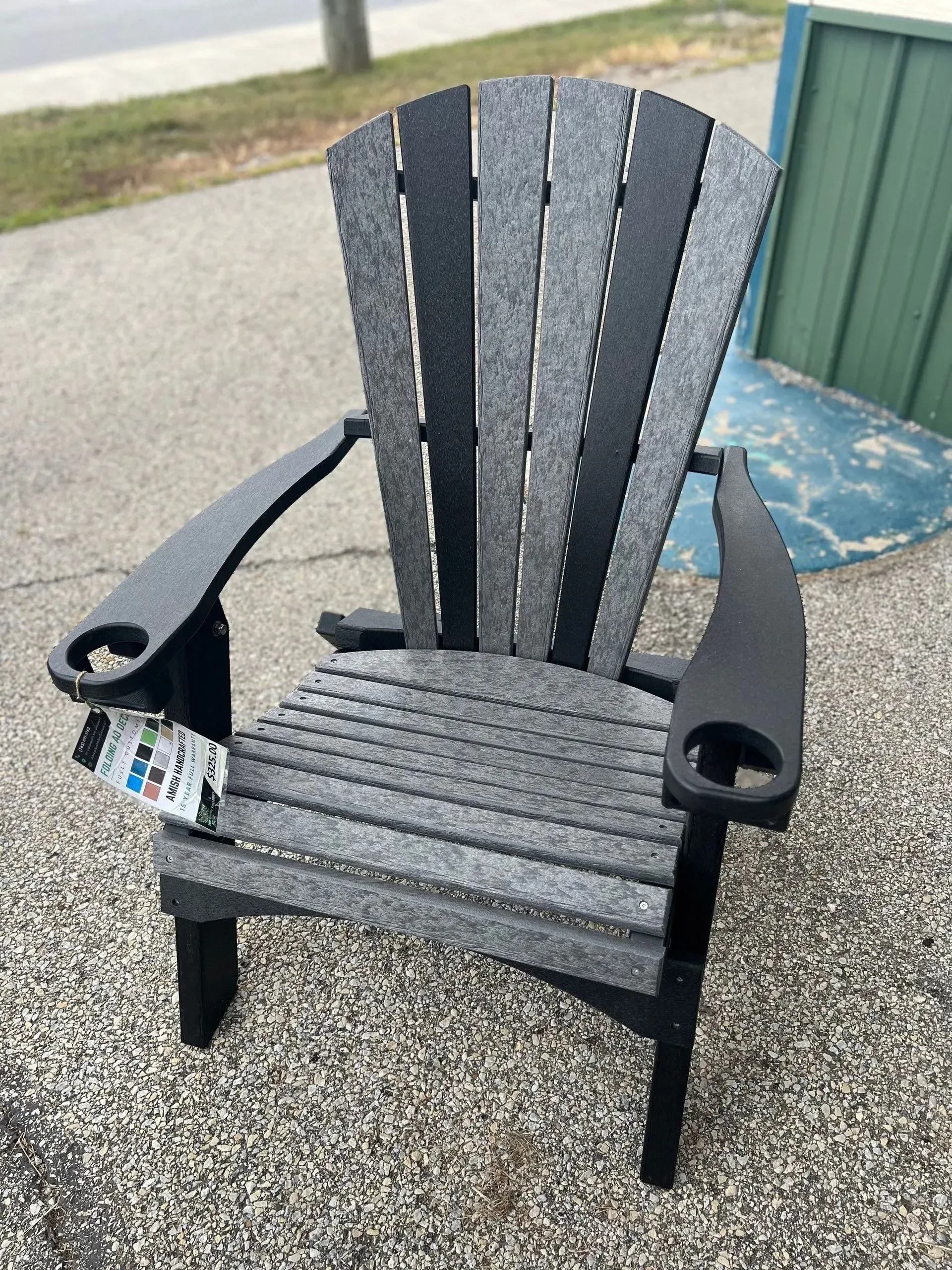 A black and gray adirondack chair is sitting on a gravel sidewalk.