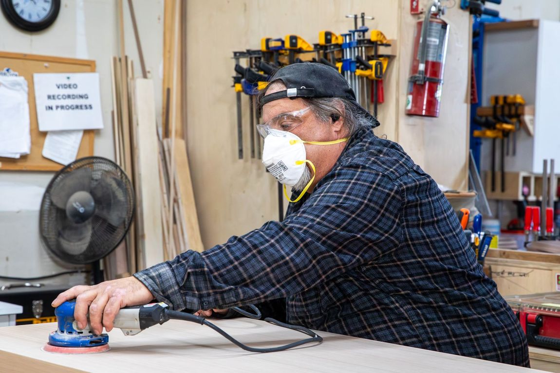 Man in a workshop sanding wood, wearing safety glasses and mask, with tools on wall.