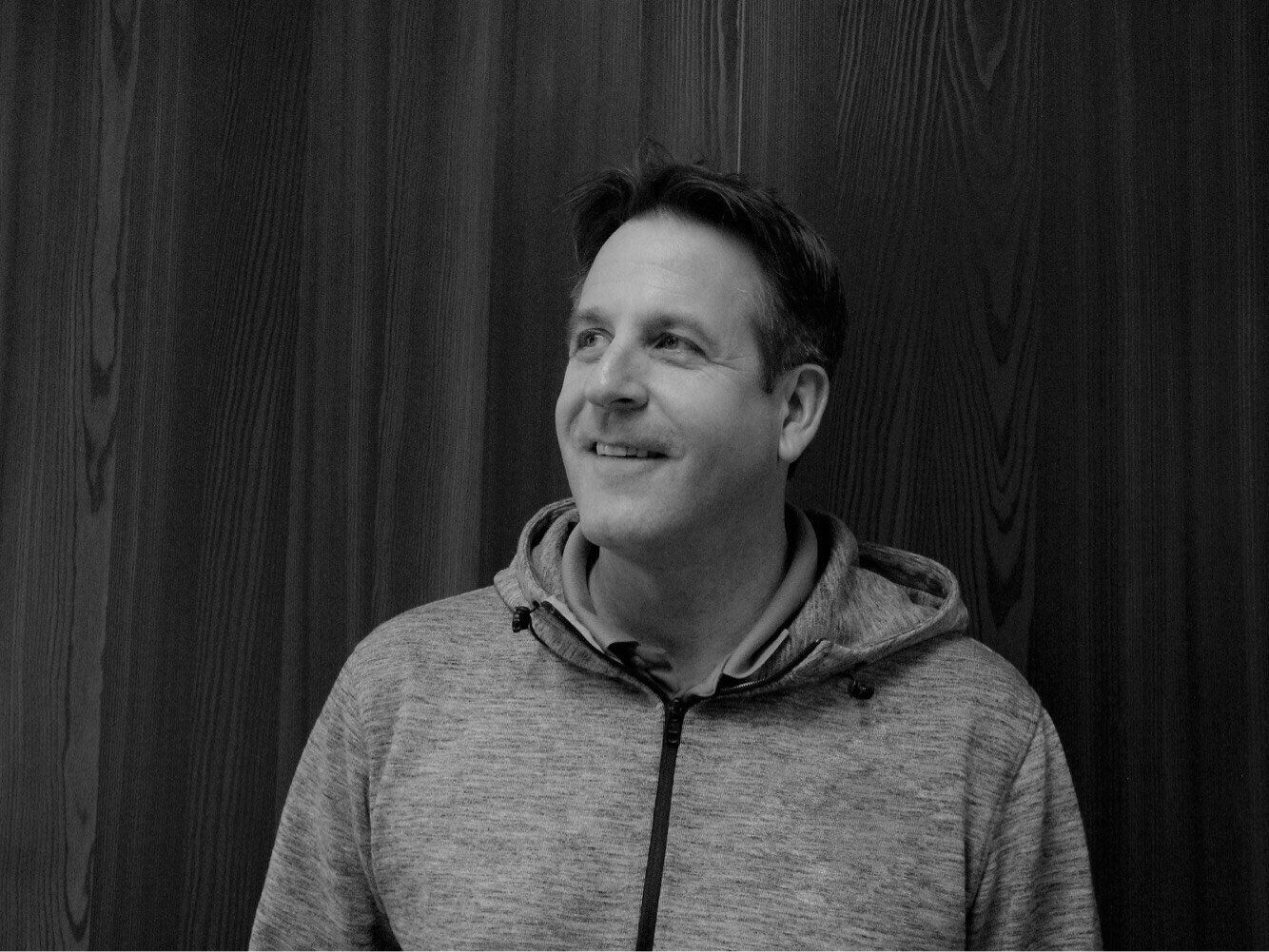 Man in a hooded sweatshirt smiles, leaning against a wood-paneled wall.