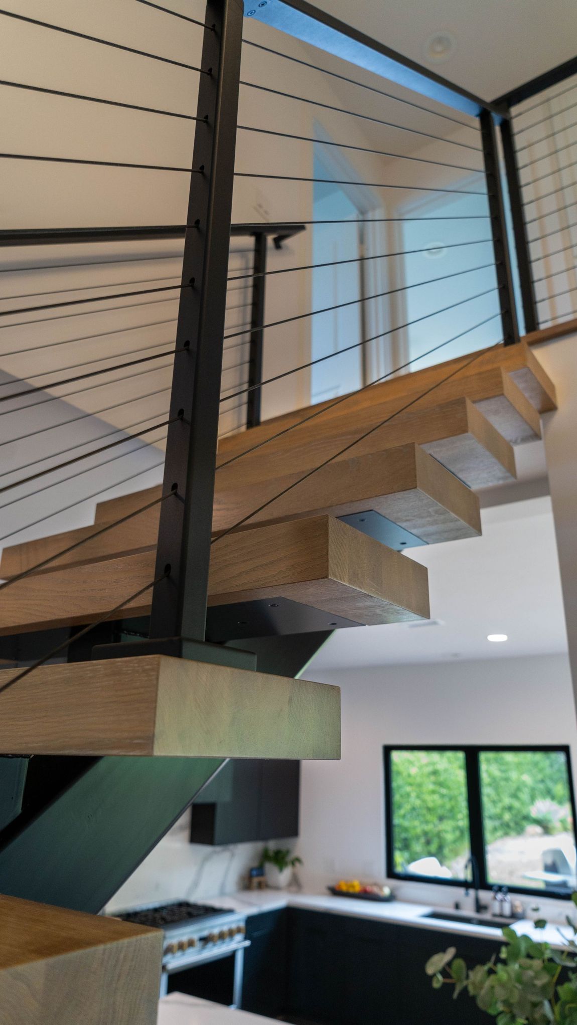Floating wooden staircase with black cable railing and dark metal posts. Kitchen visible below.