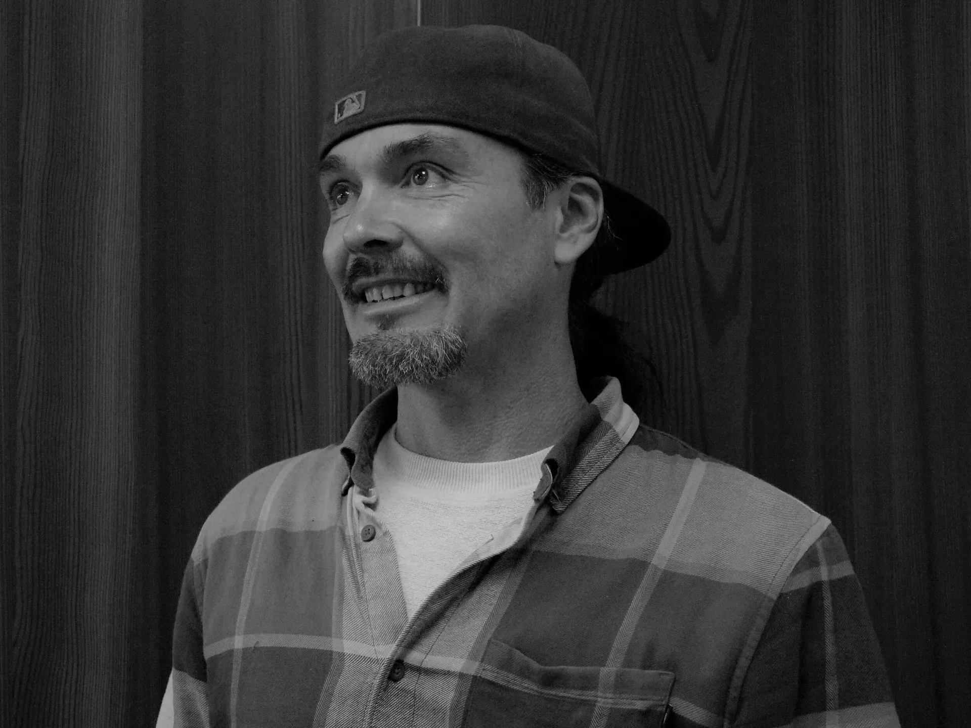 Man with beard and baseball cap smiles, wearing flannel shirt.