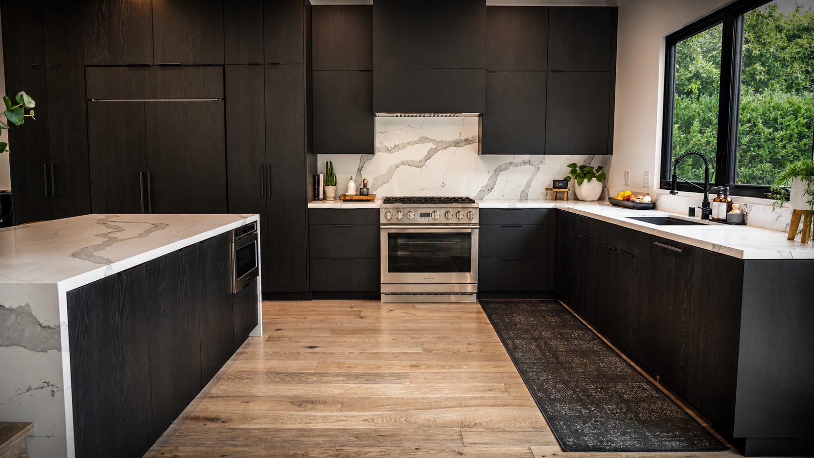 Modern kitchen with dark cabinets, marble counters, wood floor, and large window over the sink.
