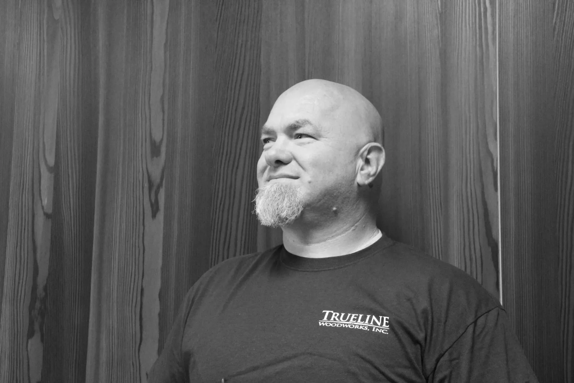 Bald man with a goatee, wearing a dark t-shirt, looking slightly to the side. Leaning against a wood-paneled wall.