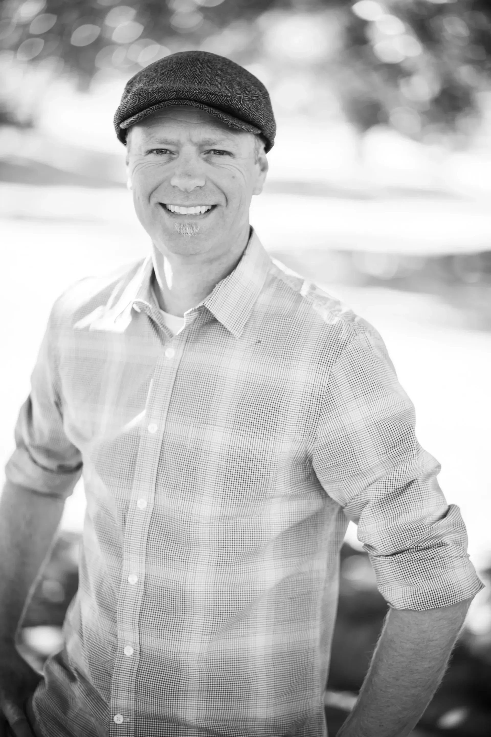 Man with a tweed cap and patterned shirt smiles outdoors, with hands on hips.