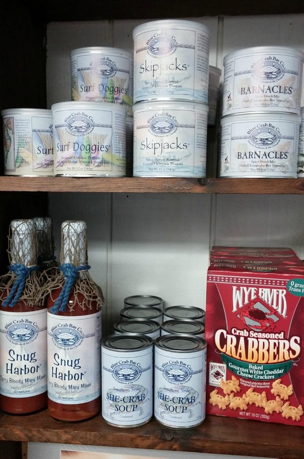 Two shelves hold various food products, including labeled tins, bottles of Snug Harbor sauce, and boxes of Wye River Crabs.