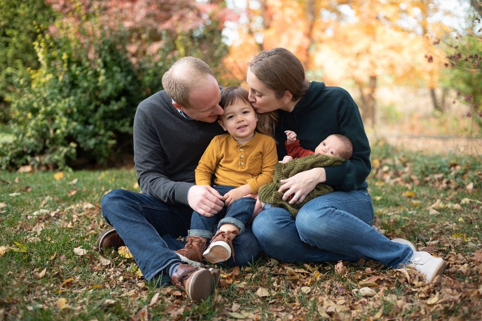Pastor and wife sitting on a lawn with their children, kissing their toddler and holding their baby