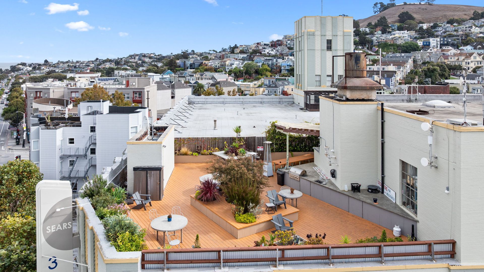 Rooftop deck with seating and plants overlooking a city with rolling hills. Sunny, daytime.