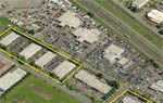 Aerial view of a commercial area with buildings, parking, and a road. Yellow outline highlights one section of buildings.