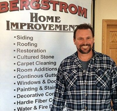 Man standing beside a Bergstrom Home Improvement sign in a plaid shirt, smiling.