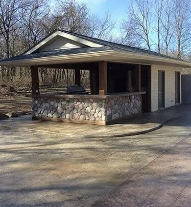 Stone pavilion with a covered roof and garage doors, standing in a wooded park area.