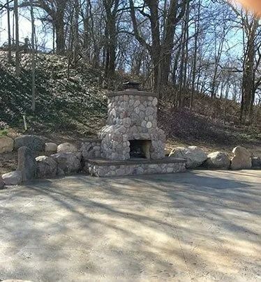 Stone outdoor fireplace in a wooded park clearing, with bare trees and a paved area in front.