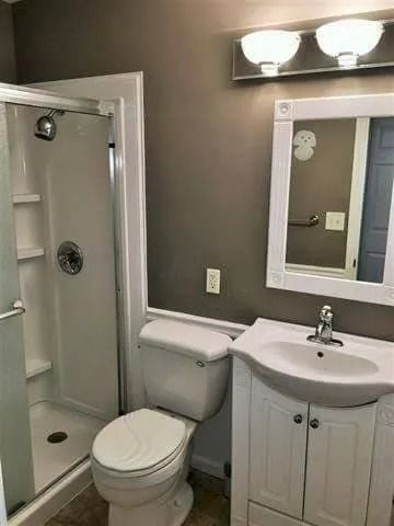 Small bathroom with toilet, sink vanity, mirror, shower, and wall light.