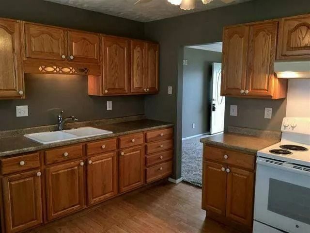 Empty kitchen with wooden cabinets, dark countertops, white stove, and a sink by a doorway.