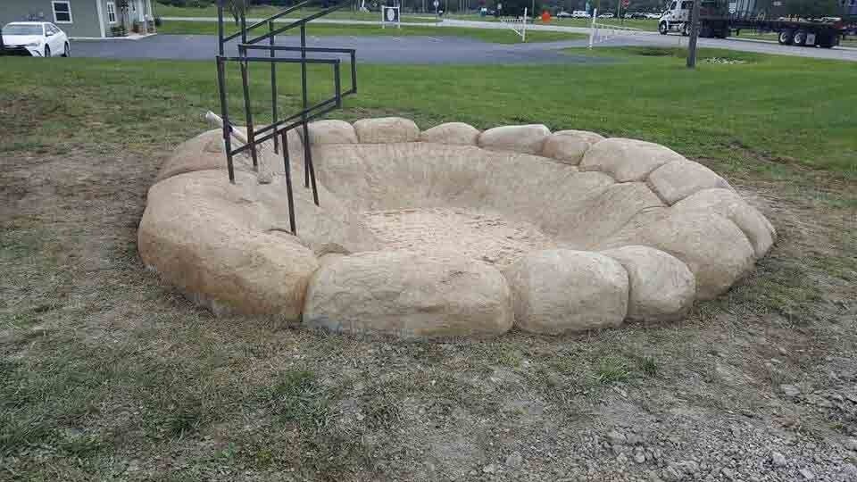 Sandbag-lined circular pit with a black handrail in a grassy outdoor area.