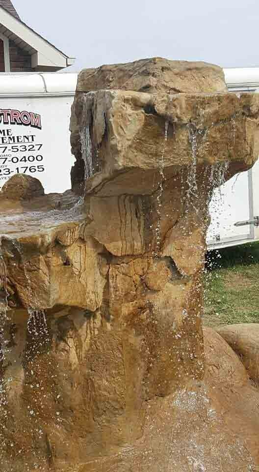 Rock waterfall fountain with water trickling over tan stone in front of a white trailer.