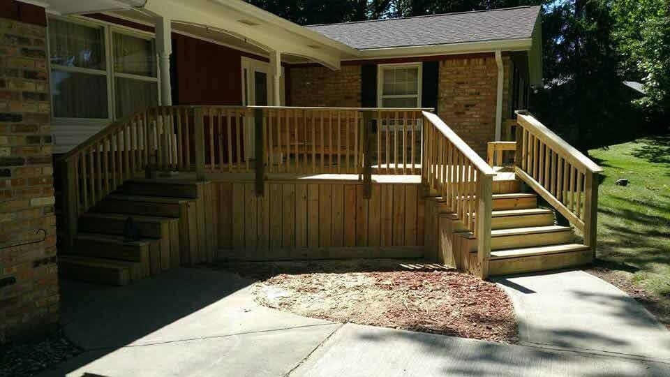 Wooden front porch with railing and steps leading to a house, beside a concrete driveway and mulch bed
