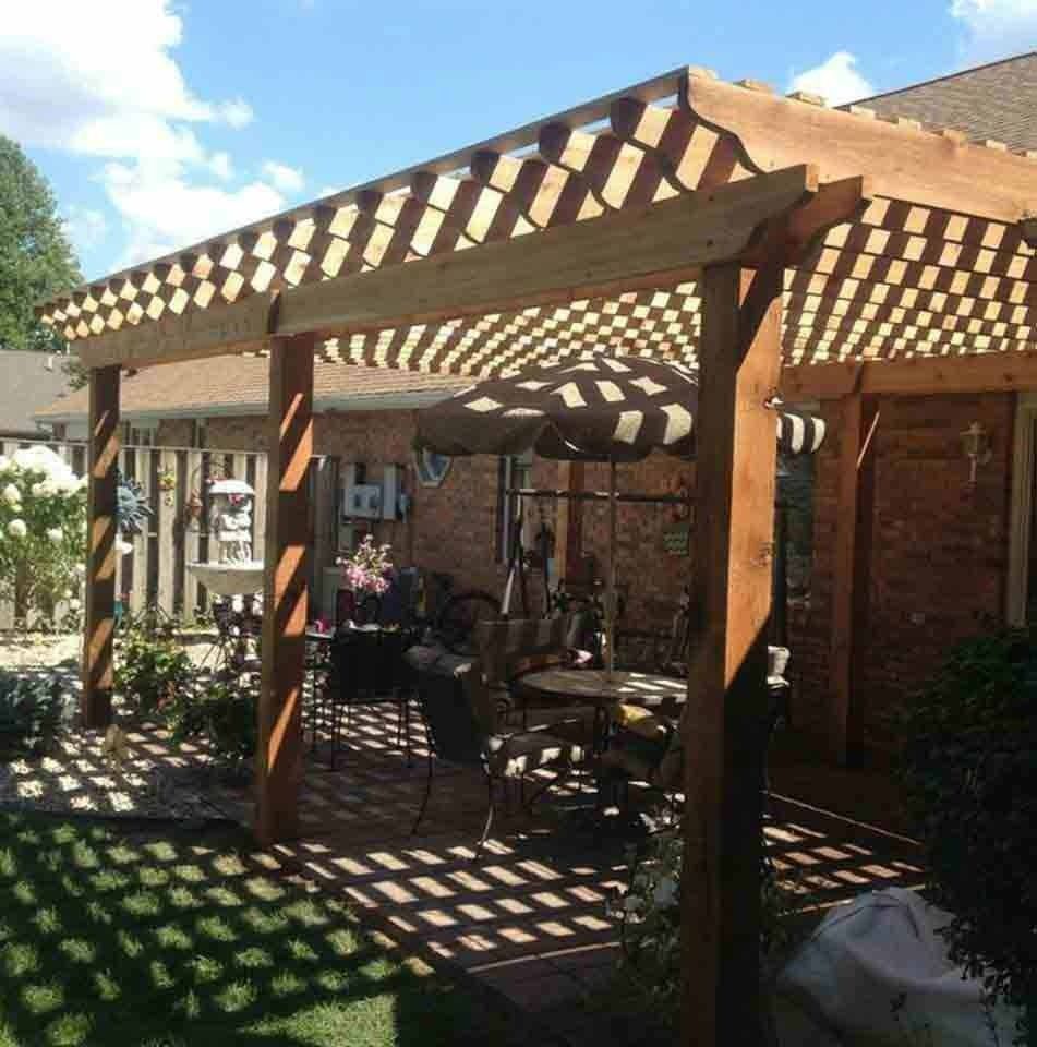 Wooden pergola with lattice roof shading a patio beside a brick house and garden, casting striped shadows
