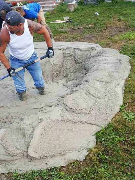 Two workers spread wet concrete over a curved outdoor surface with shovels.
