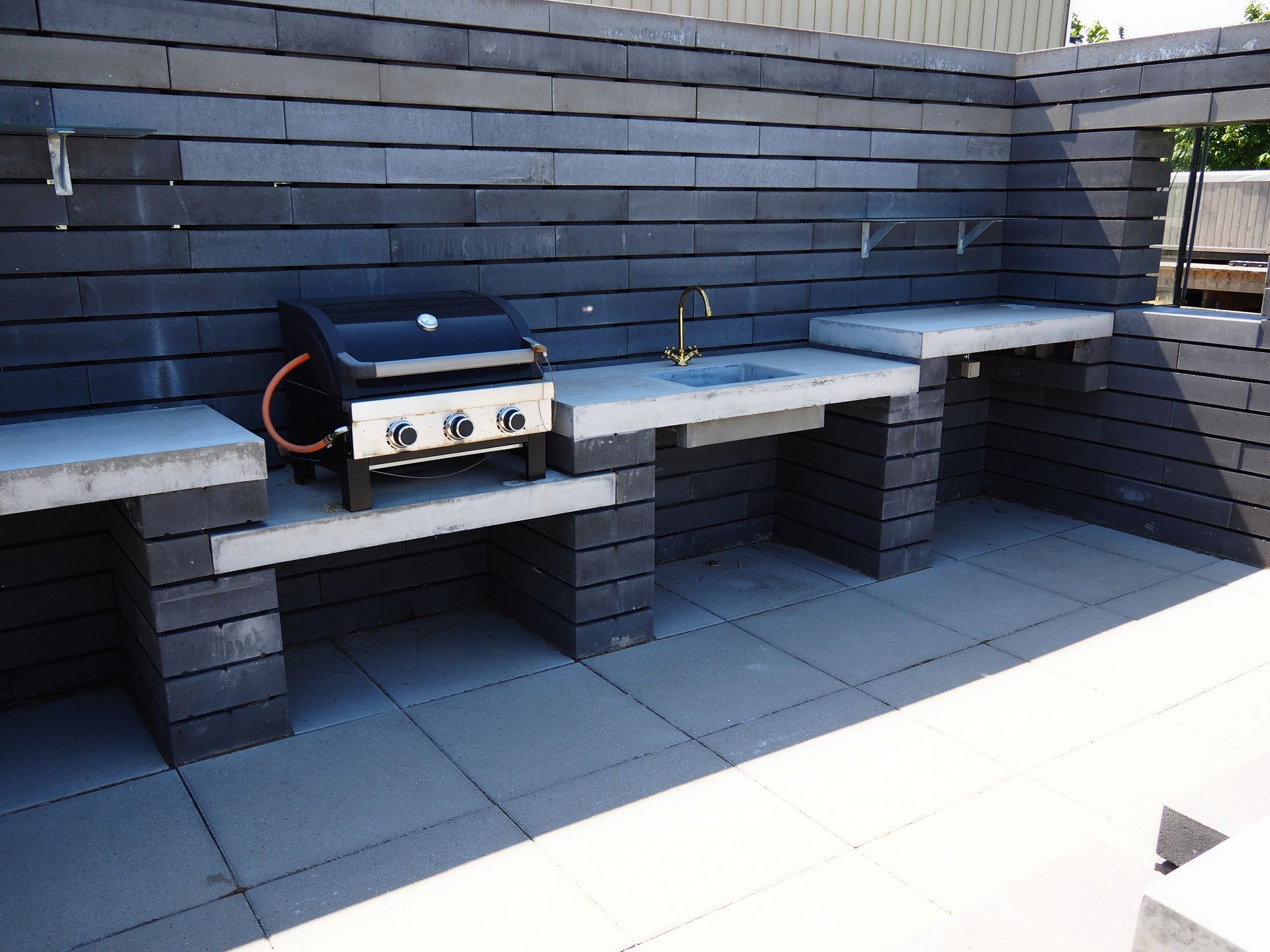 Outdoor stone barbecue area with built-in benches and a stainless steel grill against a dark brick wall