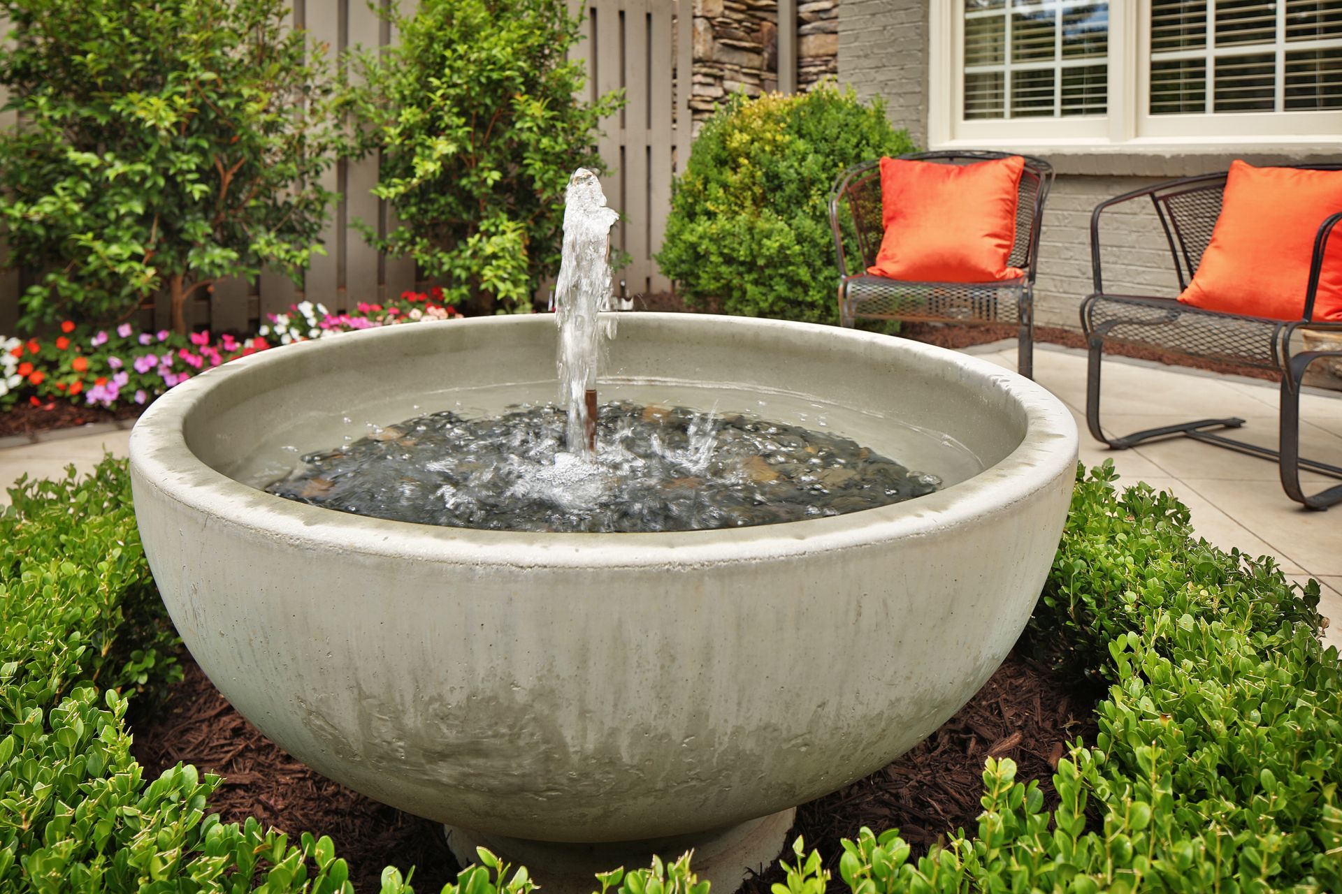 Stone fountain in a landscaped patio garden, with water spraying and orange-cushioned chairs nearby